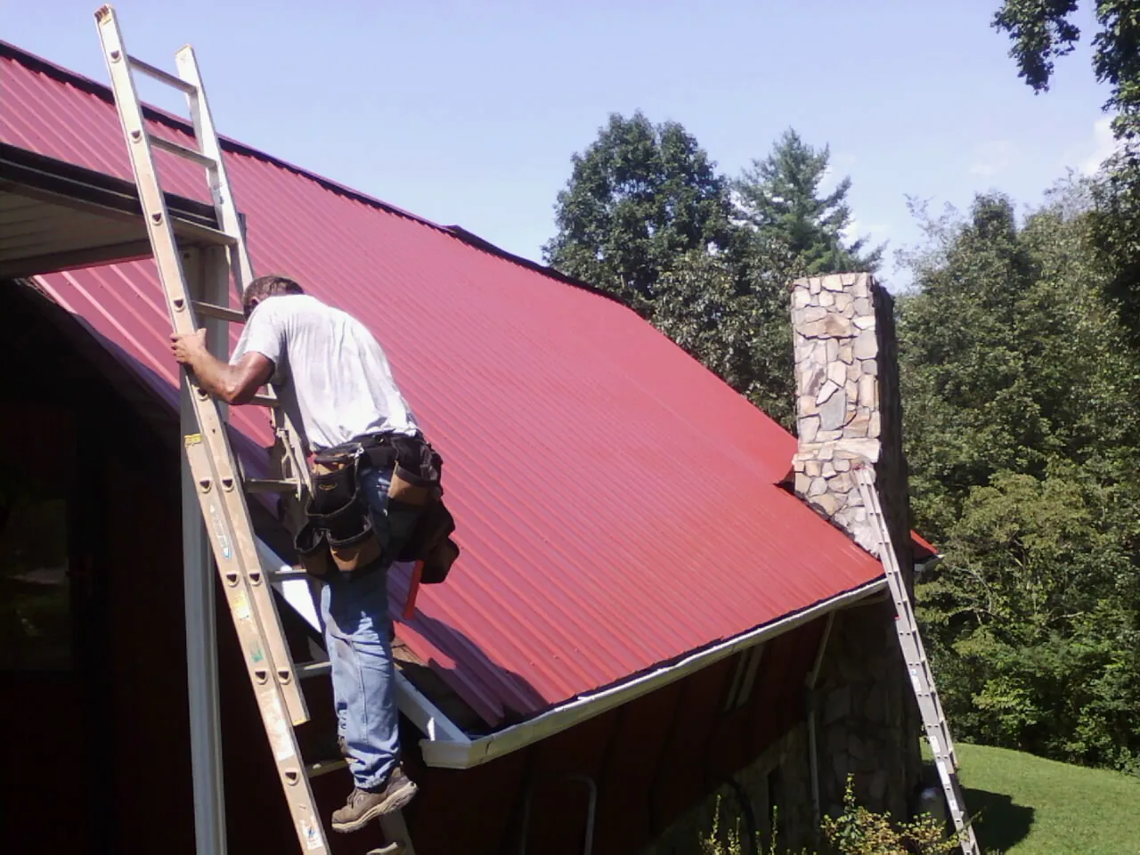 Roofer on a ladder beside a red metal roof and stone chimney — Soffit & Fascia Repair by Roofing Brothers in Dripping Springs